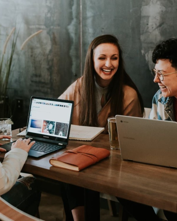Three women sit at a desk with their laptops. They are chatting and laughing.